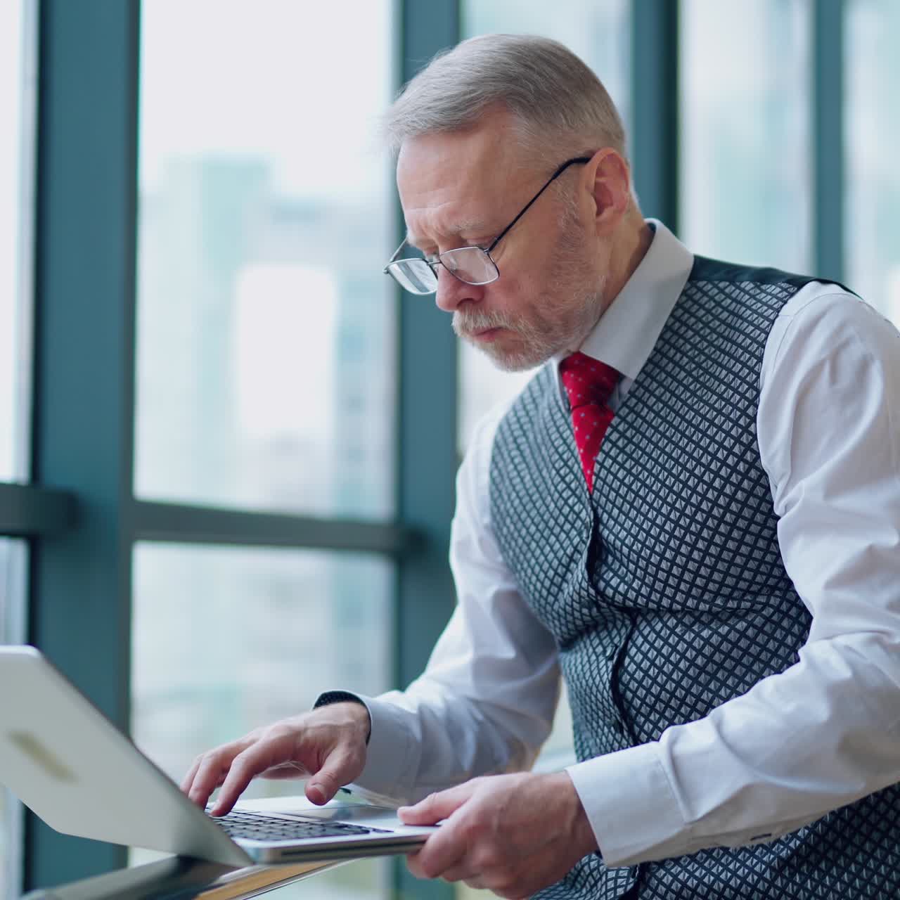 Serious businessman in glasses looking into a laptop. Mature director in elegant suit working on a laptop near the office window.