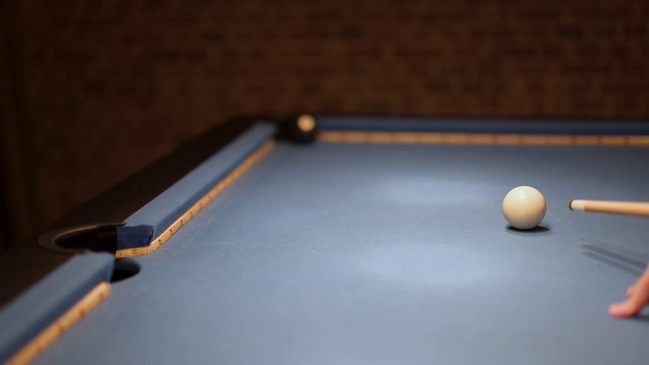 Player hits cue ball with stick on indoor billiard table, low light, brick wall background