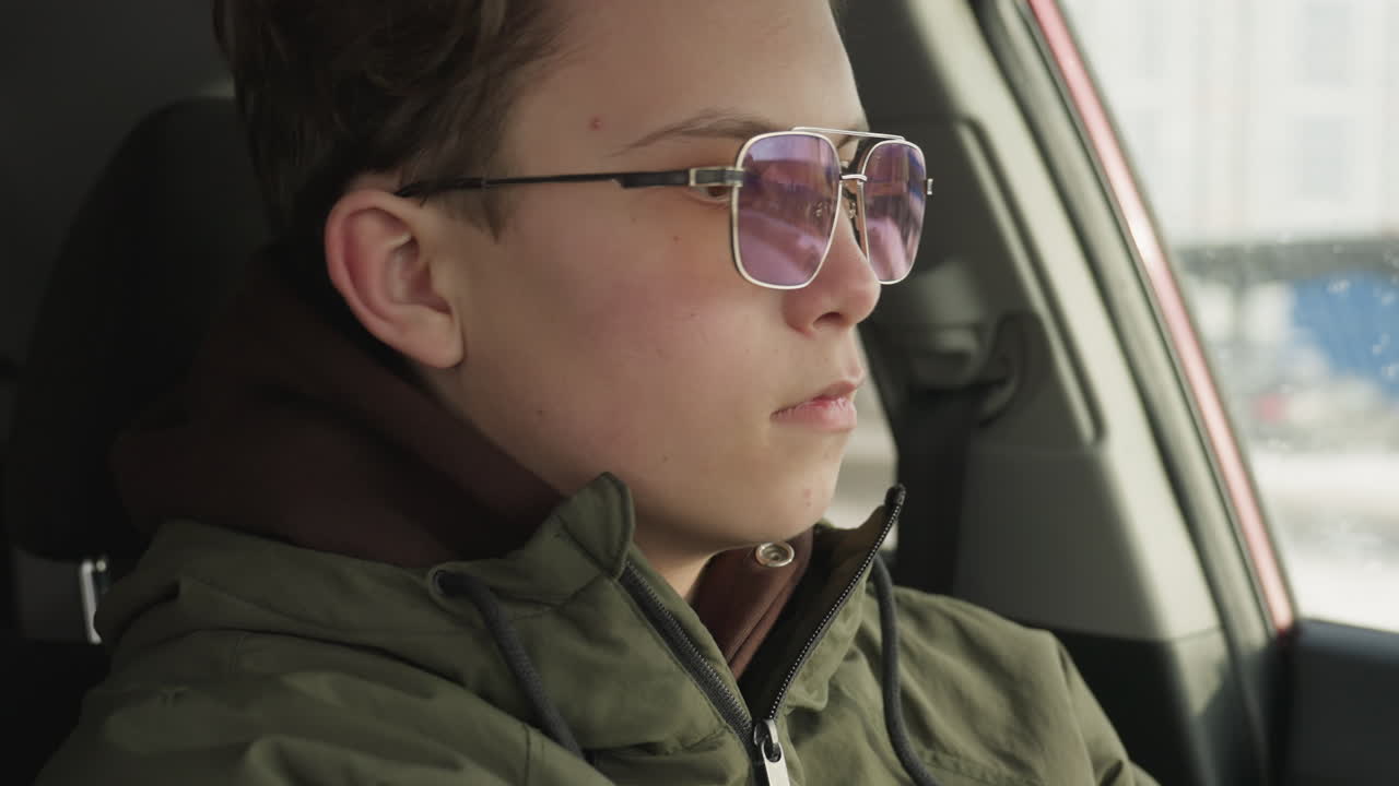 close up of young man in hoodie and glasses seated inside vehicle as he looks downward and unbuckles seatbelt during cold winter day with soft daylight entering through car window