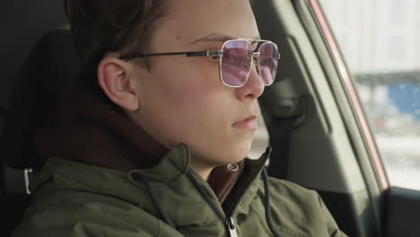 close up of young man in hoodie and glasses seated inside vehicle as he looks downward and unbuckles seatbelt during cold winter day with soft daylight entering through car window