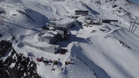 Aerial-tilt-up-shot-of-ski-resort-with-rocky-edge-and-snow-capped-mountains