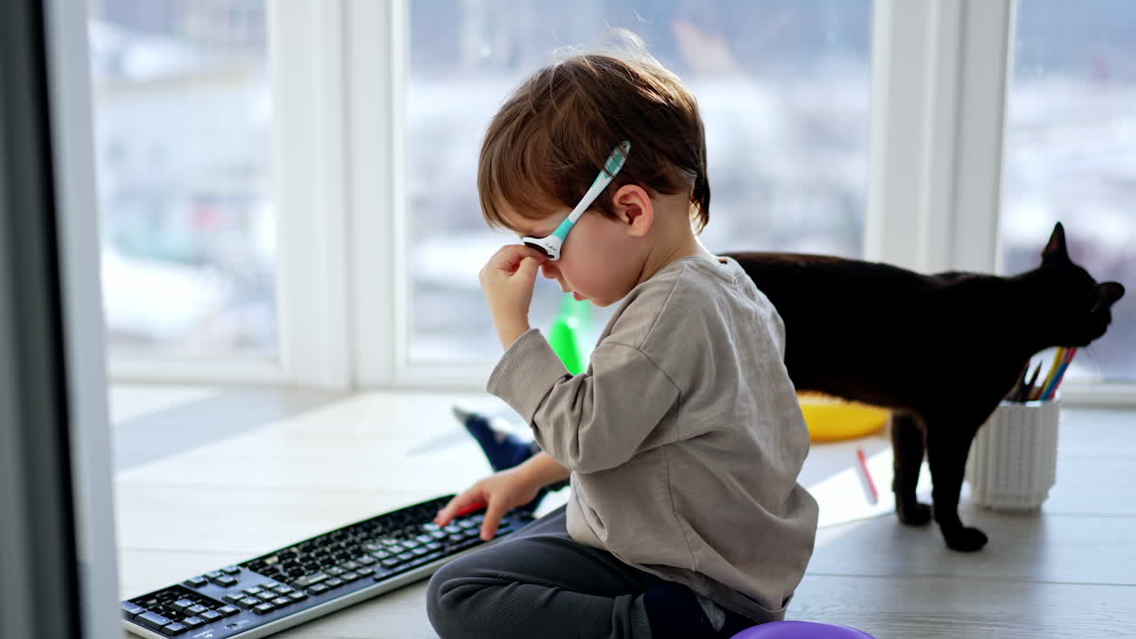 Lovely baby sitting on the floor puts on glasses. Kid takes computer keyboard and presses the keys. Black domestic cat walks around.