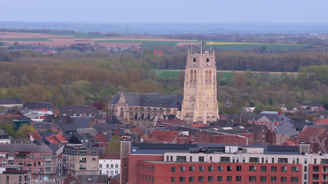 La storica torre della chiesa di Tongeren si erge sopra tetti misti e campi lontani mentre la telecamera avanza lentamente.