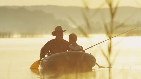 senior fisherman in hat floating with his grandson in the boat and fishing with a rod in the lake