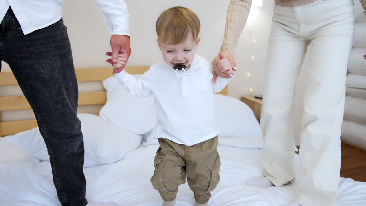 Unrecognized parents and lovely little baby with pacifier stand on the bed. Happy family play together at home.