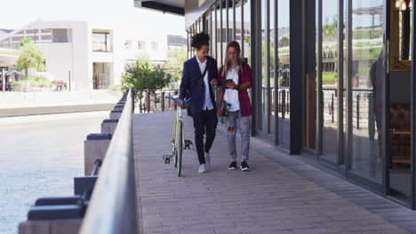 Two-diverse-male-friends-walking-in-the-street-using-a-digital-tablet-and-wheeling-a-bicycle