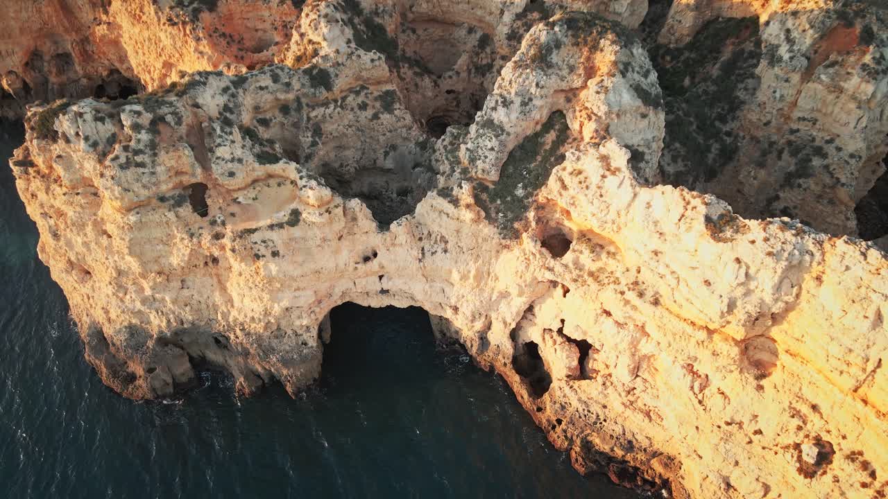 Camera moves forward, sweeping across sunlit limestone cliffs and arches, and gliding toward Ponta da Piedade lighthouse, with the coastline and town on the horizon
