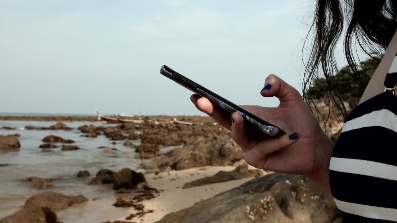 소녀는 바다와 돌의 배경에서 해변에서 휴대전화를 사용합니다. 메시지를 쓴다.