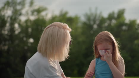 young girl wiping tears with tissue outdoors, sad expression with glistening eyes, soft sunlight and blurred green background creating quiet emotional moment of vulnerability and reflection