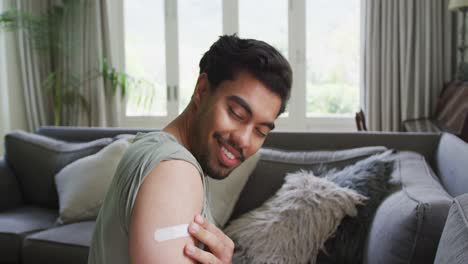 Portrait-of-biracial-young-man-with-bandage-on-arm-sitting-at-home-after-immunity-vaccination