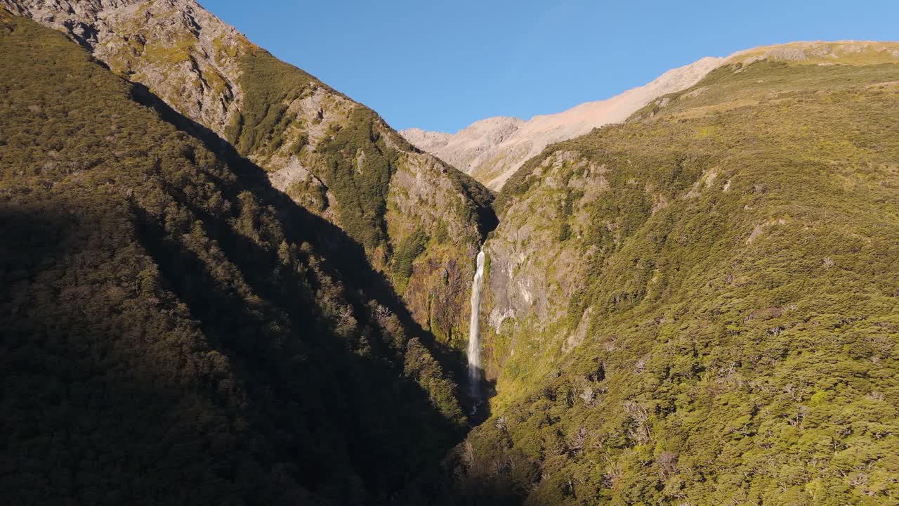 Gigantic Devil’s Punchbowl Waterfall at Arthurs Pass during sunset time in New Zealand. Aerial approaching shot. Blue sky in summer. Wide shot.