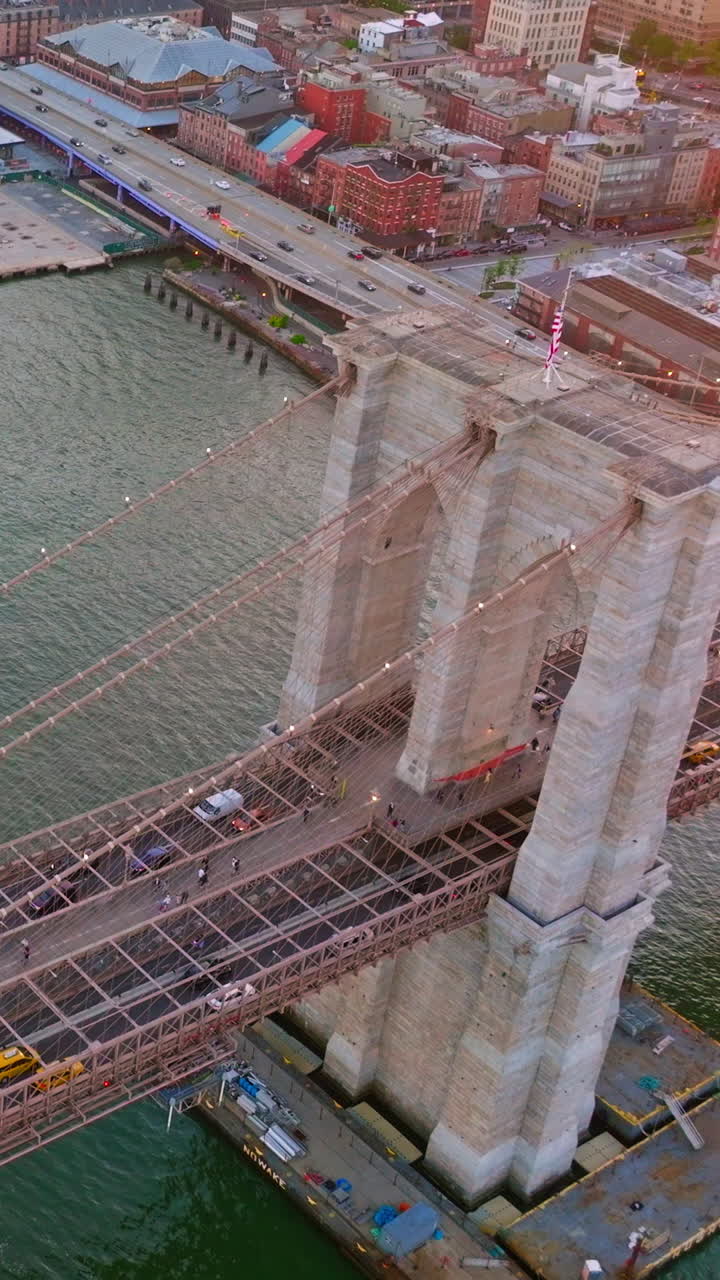 Famous Brooklyn Bridge over East River in New York. Numerous cars going along the bridge and yacht coming under it. Aerial view. Vertical video