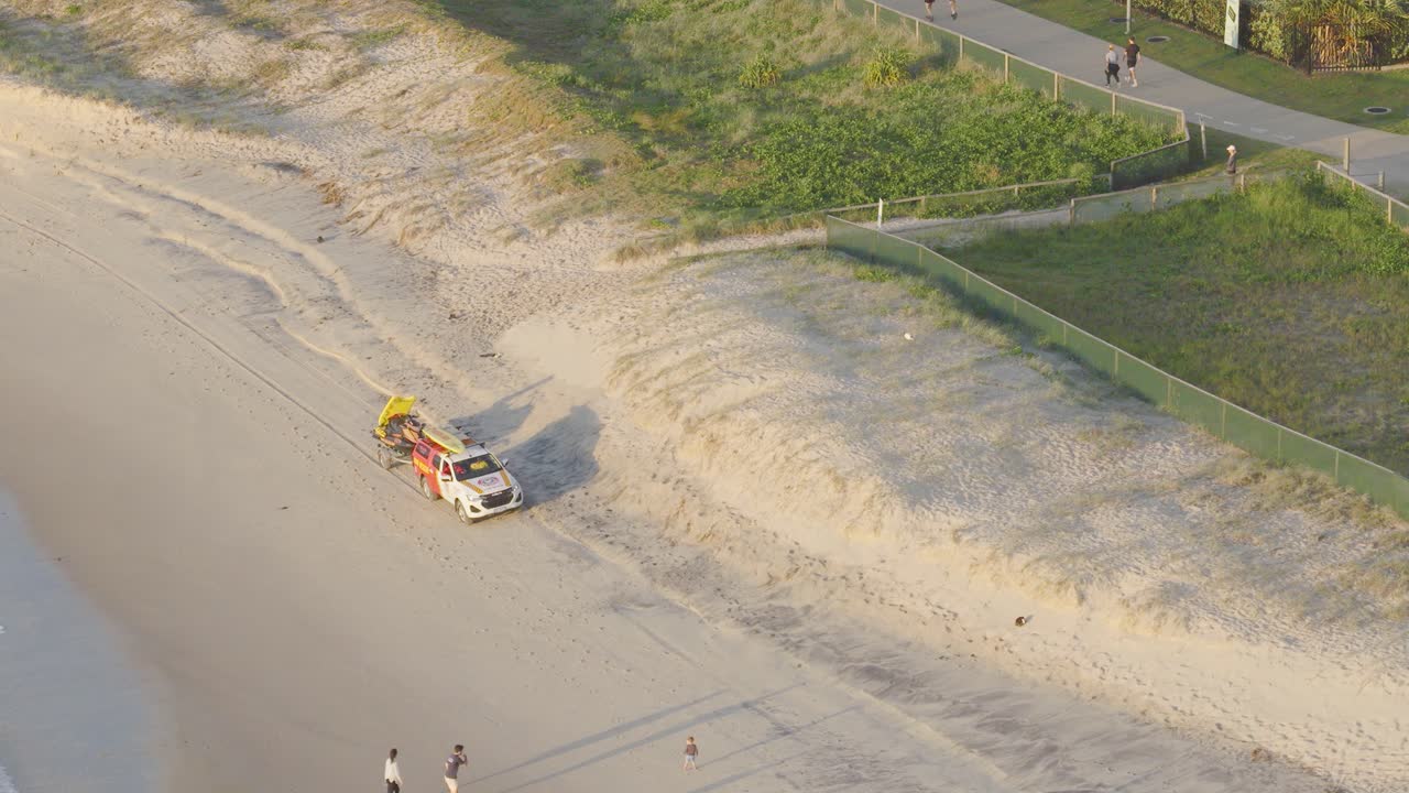 Aerial view of a patrol vehicle driving along Gold Coast beach, with people walking nearby under warm sunlight