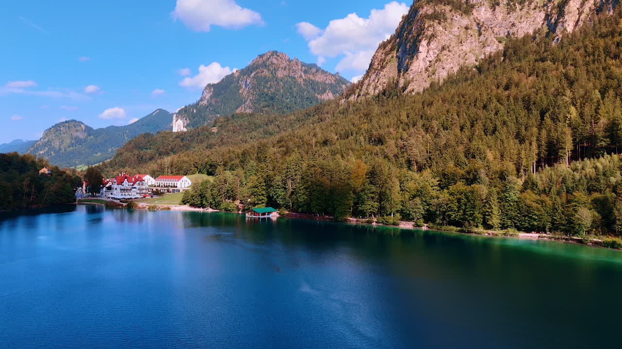 Flight over the beautiful lake surrounded by the mountains covered with pine tree woods. Lovely white houses with red roofs at the bank. Scenery of Bavaria, Germany
