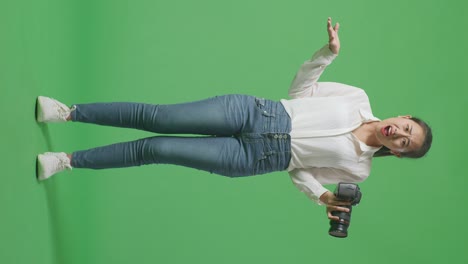 cuerpo lleno de fotógrafo asiático desconcertado usando una cámara tomando fotos, diciendo por qué y haciendo gestos dudosos mientras está de pie en el fondo de la pantalla verde en el estudio