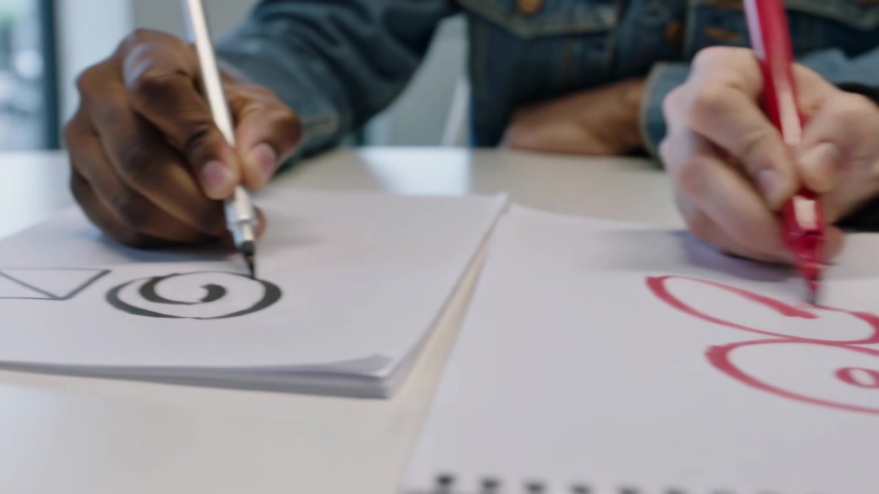 Colleghi che iniziano tratti a sinistra, si esercitano a disegnare su blocchi da tavolo bianchi usando un pennello nero e una penna rossa, indossando una felpa con cappuccio.