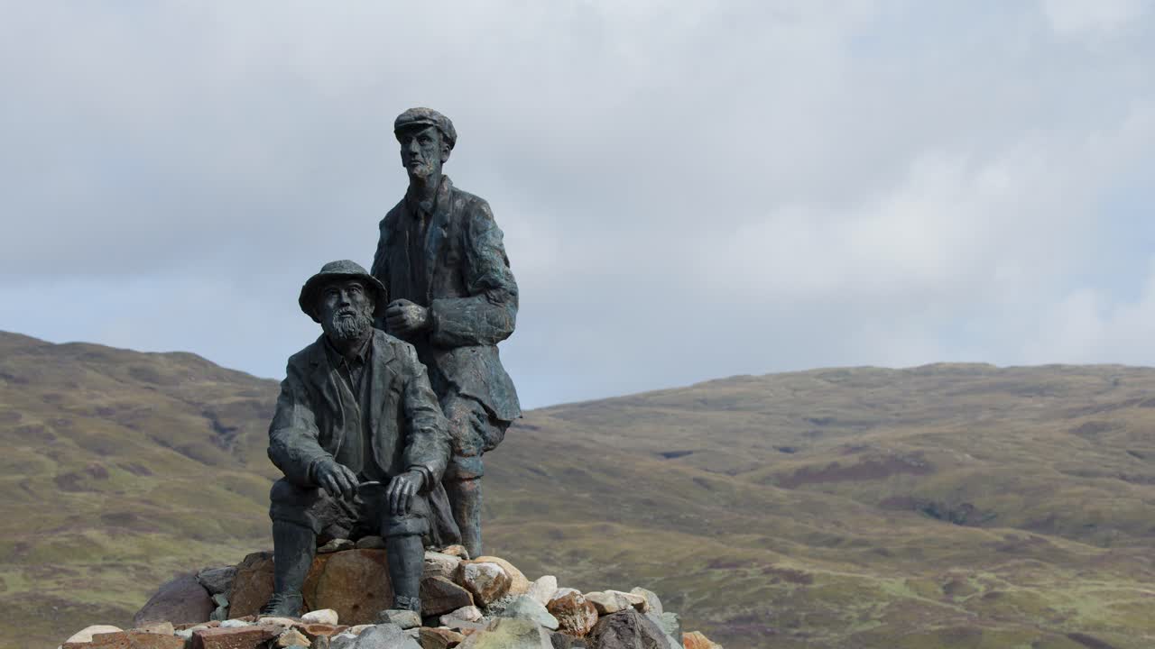 Bronze sculpture of two men on rocky base, static wide shot, overcast daylight, mountainous background