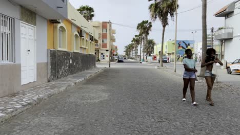 girls walking along tranquil street in sal rei, boa vista, cape verde, lined with palm trees and colorful buildings, capturing the essence of island life and local architecture