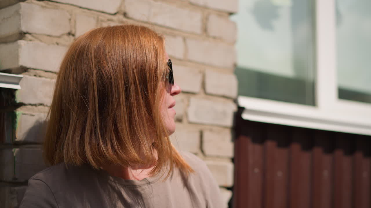 Woman with light brown hair lean against sunlit wall during warm afternoon, adjusting sunglasses while turning head right and left, sunlight creating gentle highlights