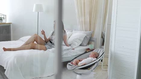 mom looks at the mobile phone and writes to social networks, and next to the baby caries chair in a white interior