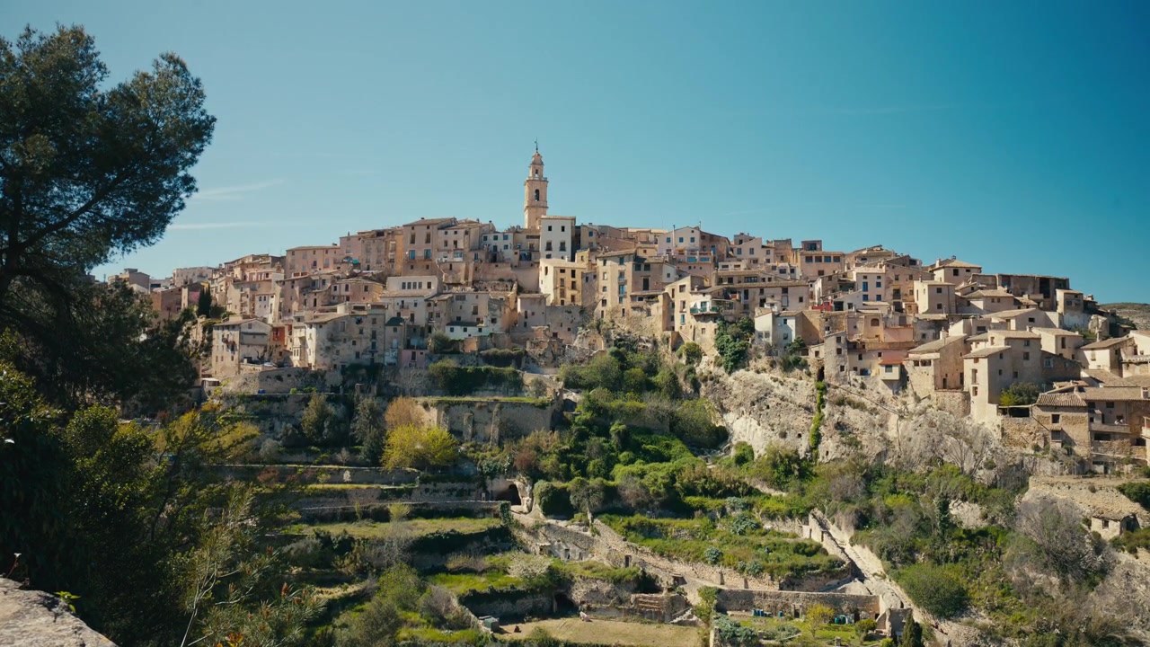 Una veduta panoramica rivela un borgo medievale collinare che si erge sopra terrazzamenti verdi, con un campanile, case in pietra sovrapposte e un tranquillo paesaggio mediterraneo a Bocairent, Spagna