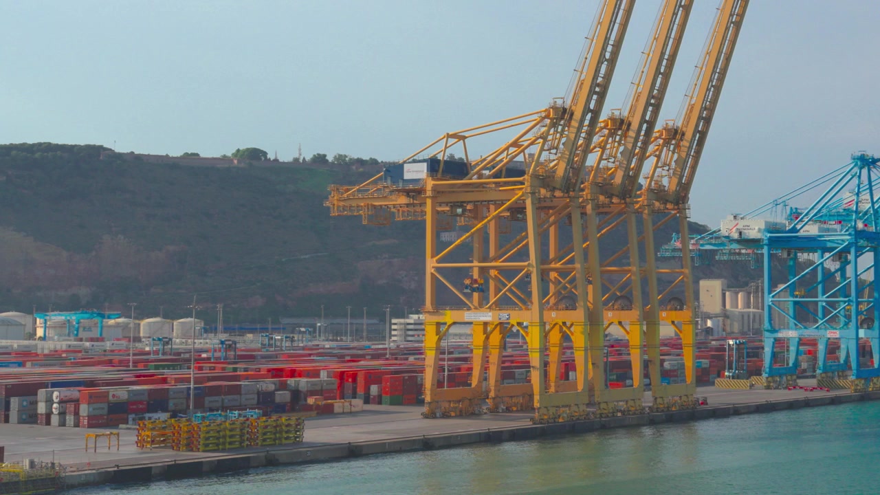 Large gantry cranes moving along the quayside in a busy commercial harbor, with shipping containers stacked in the background, representing global logistics and international trade operations