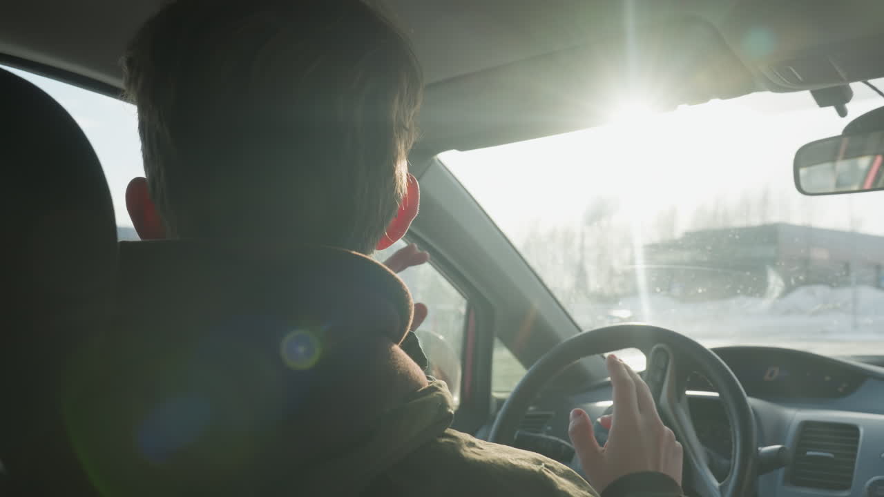 man in warm hoodie dancing inside car with hands lifted, sunlight shining through windshield creating bright flare and silhouette effect, serene winter background with trees and blurred scene
