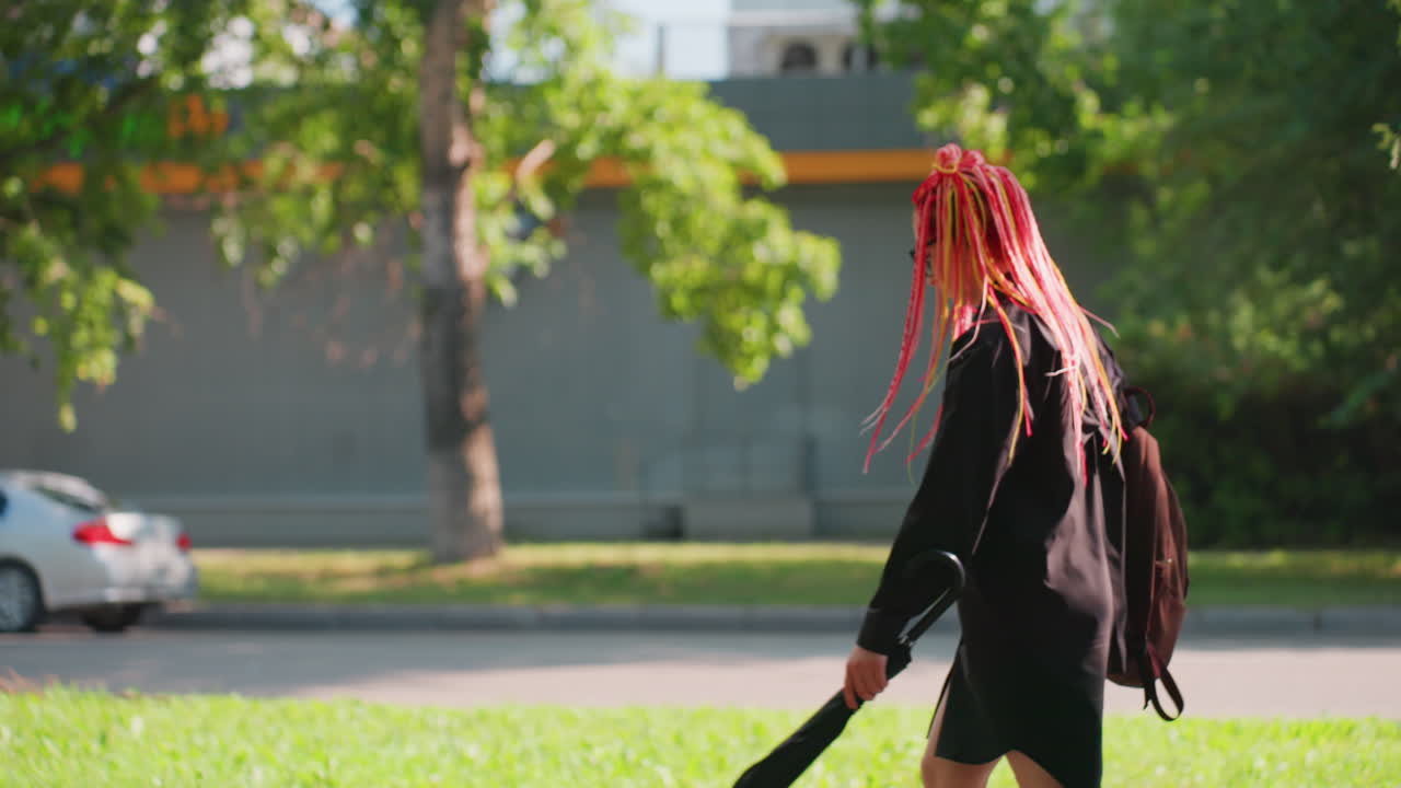 흐릿한 인물 풍경, 흐릿하게 멀리 보이는 사람들과 길가의 풍경이 있는 전경의 나무, 전경의 나무 줄기가 분홍색 땋은 머리와 우산을 든 흐릿한 배경 속 인물을 포착합니다.