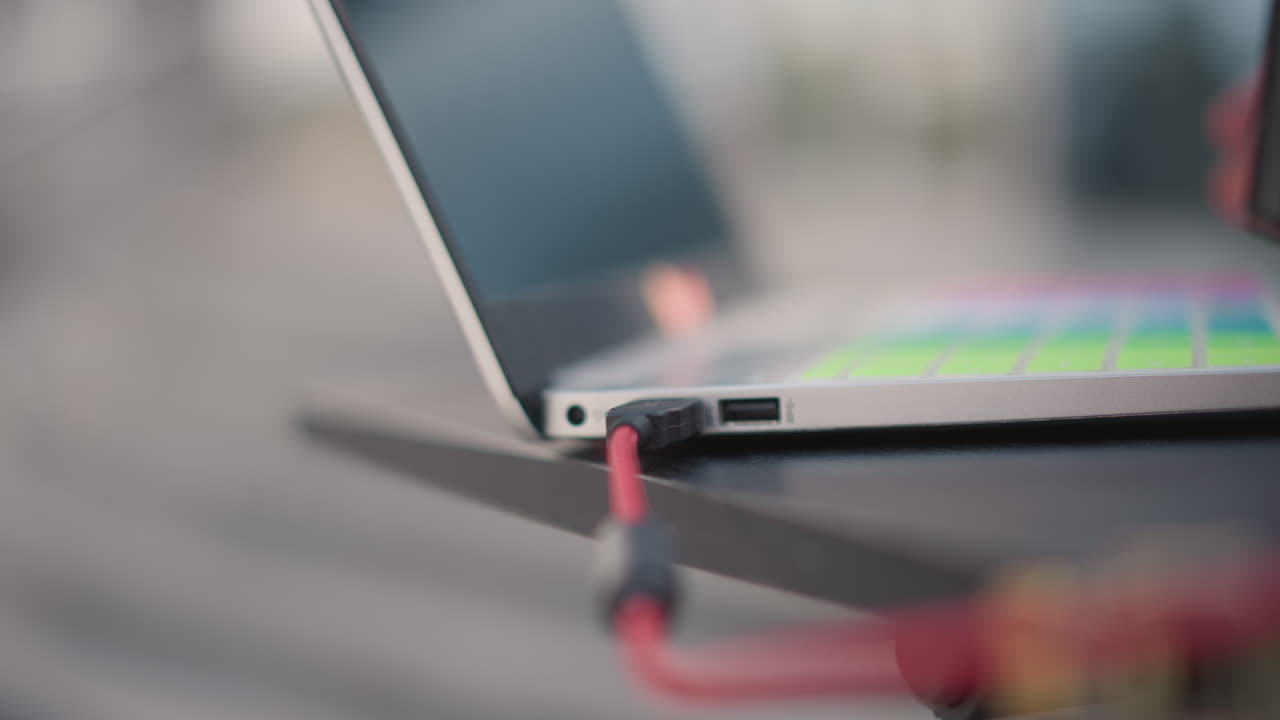 laptop edge with red cable plugged, closeup of dj laptop on compact stand showing vibrant backlit keyboard, hand about to secure braided cord, urban outdoor energy, anticipation before set