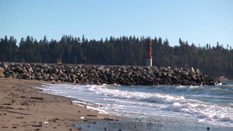 Junge-Frau,-Die-An-Einem-Sonnigen-Frühlingstag-Von-Einer-Hohen-Statue-Auf-Einem-Felsigen-Pier-Am-Strand-Weggeht,-Wobei-Die-Wellen-Das-Ufer-Treffen