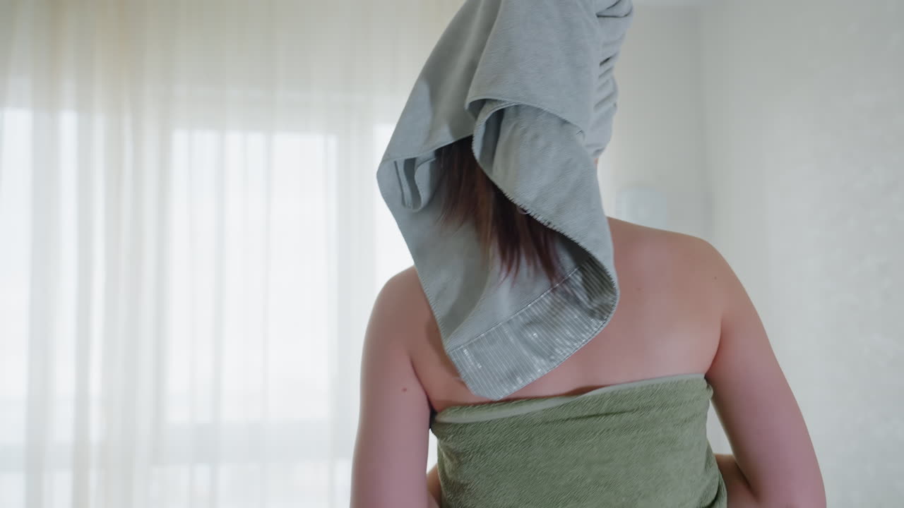 Side view of woman with hair wrapped in towel and green towel around body holding nail file at chest while sitting on sofa in living room with sheer curtains chandelier overhead