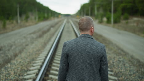 a back view of an aged man in a gray jacket, walking alone on a railway track, with a blurred view of trees and electric poles around