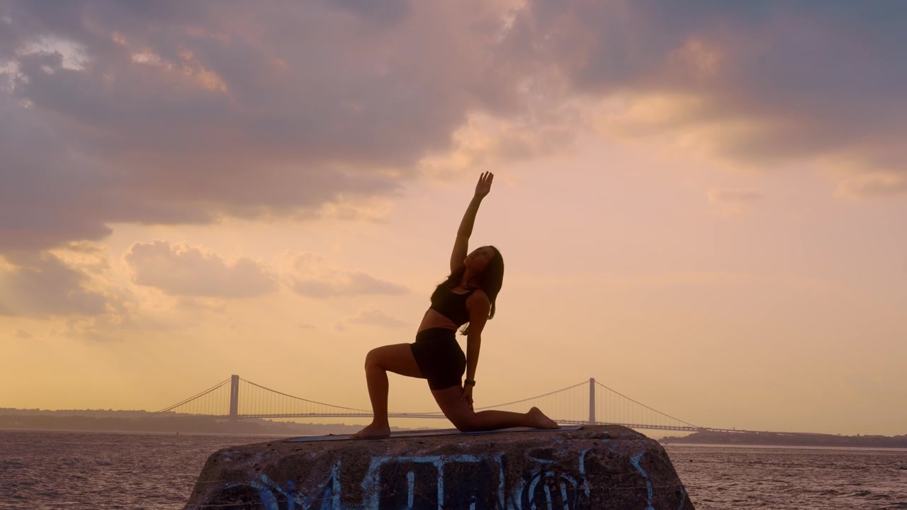 Donna che pratica la posizione yoga dell'affondo a mezzaluna al tramonto a New York. Silhouette di una persona su un molo di cemento contro il ponte Verrazzano Narrows e un cielo dorato.