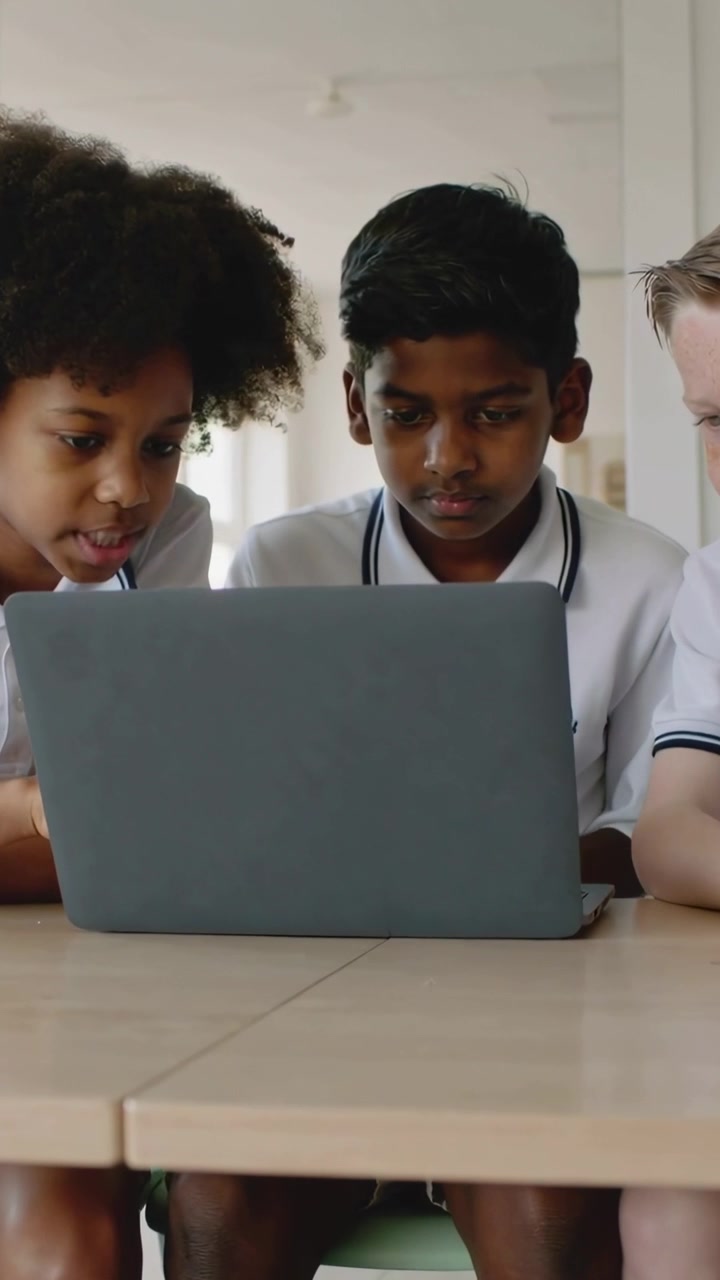 Vertical video: Leaning left pupil in white polos guiding peers, studying gray laptop in classroom
