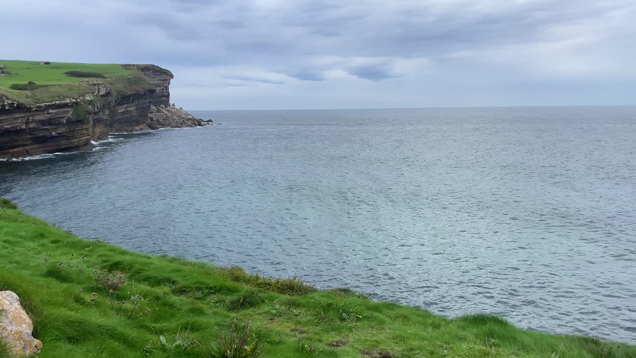 Beautiful shot of the Cantabrian Sea where we see the sea that is calm and how it hits the coast and we appreciate the horizontal strata on the cliff wall, there is a blanket of grass on top.