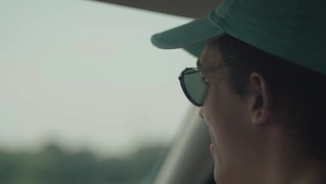 [close up] shot from the rear of a car of a young man wearing a blue cap and a man bun singing and moving his head to music