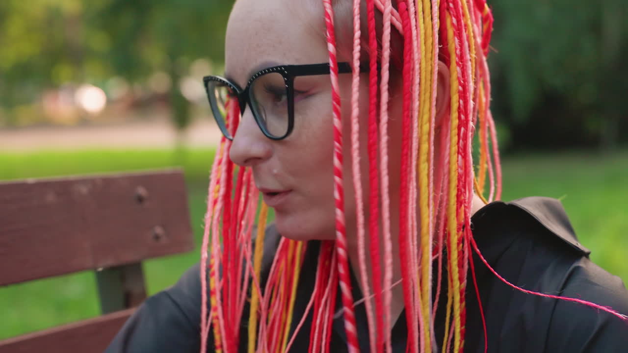 Un joven mantiene la compostura mientras se arregla un peinado colorido al aire libre; una persona sentada al aire libre muestra una energía inquieta mientras se ajusta cuidadosamente su vibrante peinado con nerviosa concentración.