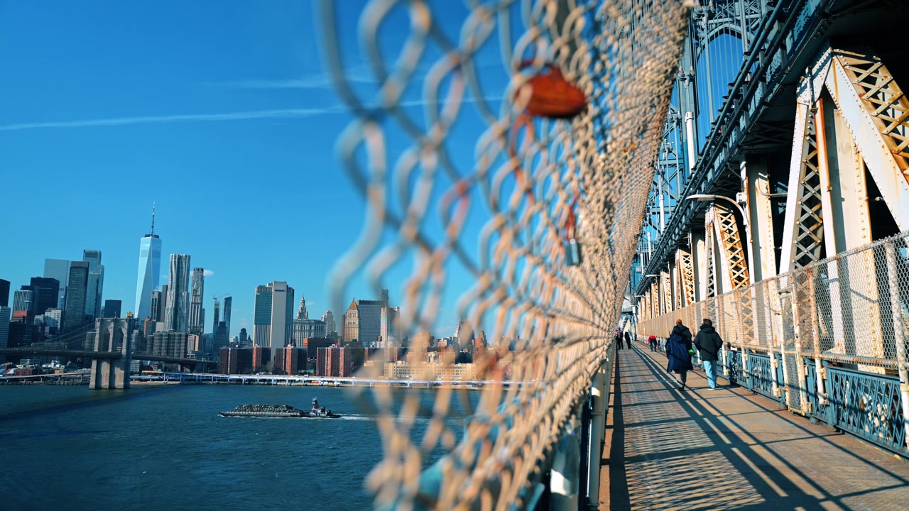 Pedoni che camminano sul Ponte di Manhattan sopra l'East River. Un rimorchiatore traina una chiatta sotto la passerella mentre lo skyline di Manhattan si estende lungo l'orizzonte.