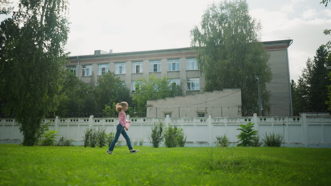 Side view of little girl walking casually across green lawn while swinging pink balloon, with tall trees and multi-story urban building in background under soft daylight