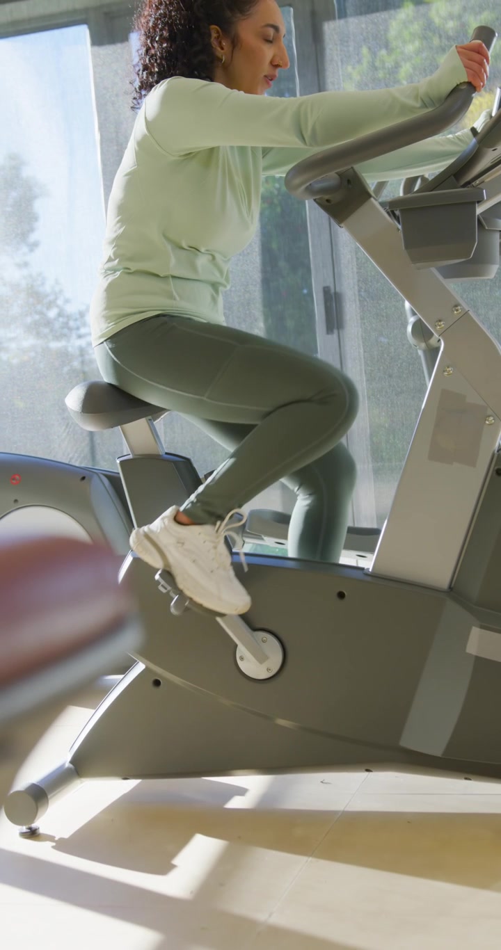 Video verticale: Donna adulta vestita di verde che si sistema su una cyclette in palestra, pedalando per fare cardio, controllando il telefono.