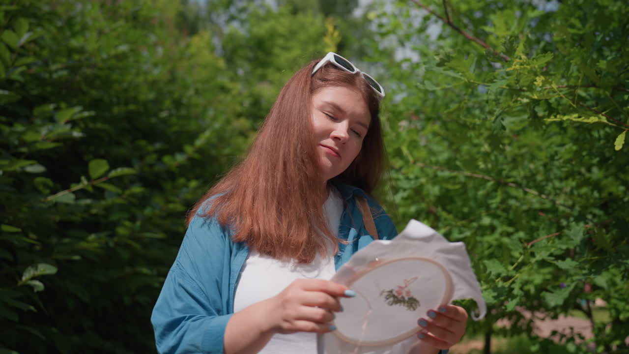 Chubby woman wearing blue shirt and white outfit walking along sunny green alley holding embroidered fabric, dancing joyfully under leafy trees, enjoying creative inspiration