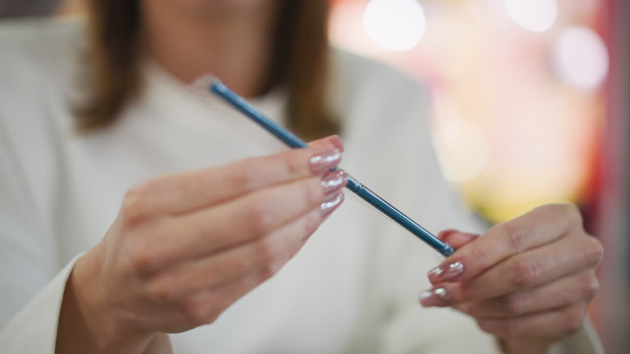 primer plano de manos con uñas pintadas sosteniendo una paja en un envoltorio transparente, examinándola cuidadosamente en un entorno interior moderno con un fondo vibrante borroso e iluminación suave