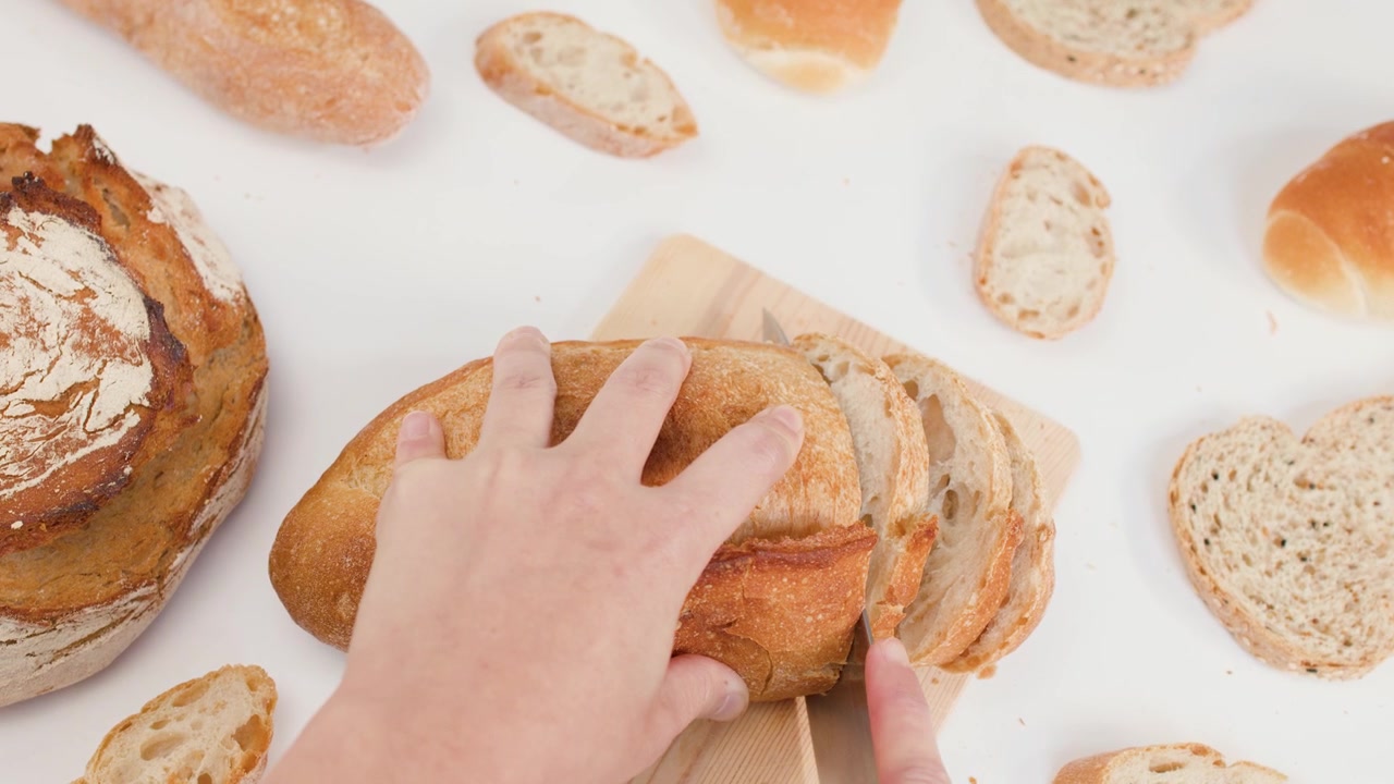 Top-down view of hands using a knife to slice crusty sourdough bread on white surface.