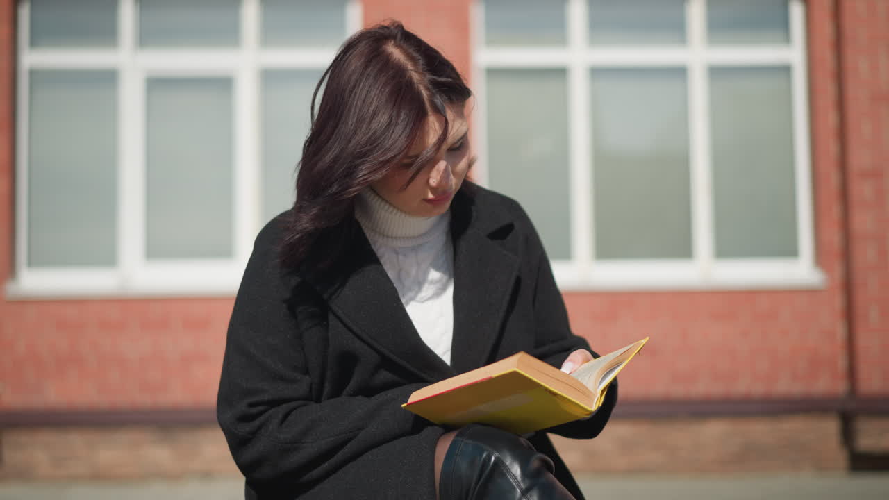 Woman in black coat sits outside, focused on reading her book, sunlight gently illuminates her as she turns the page, flower pots sway in the breeze, and a lamp post casts a shadow on the ground