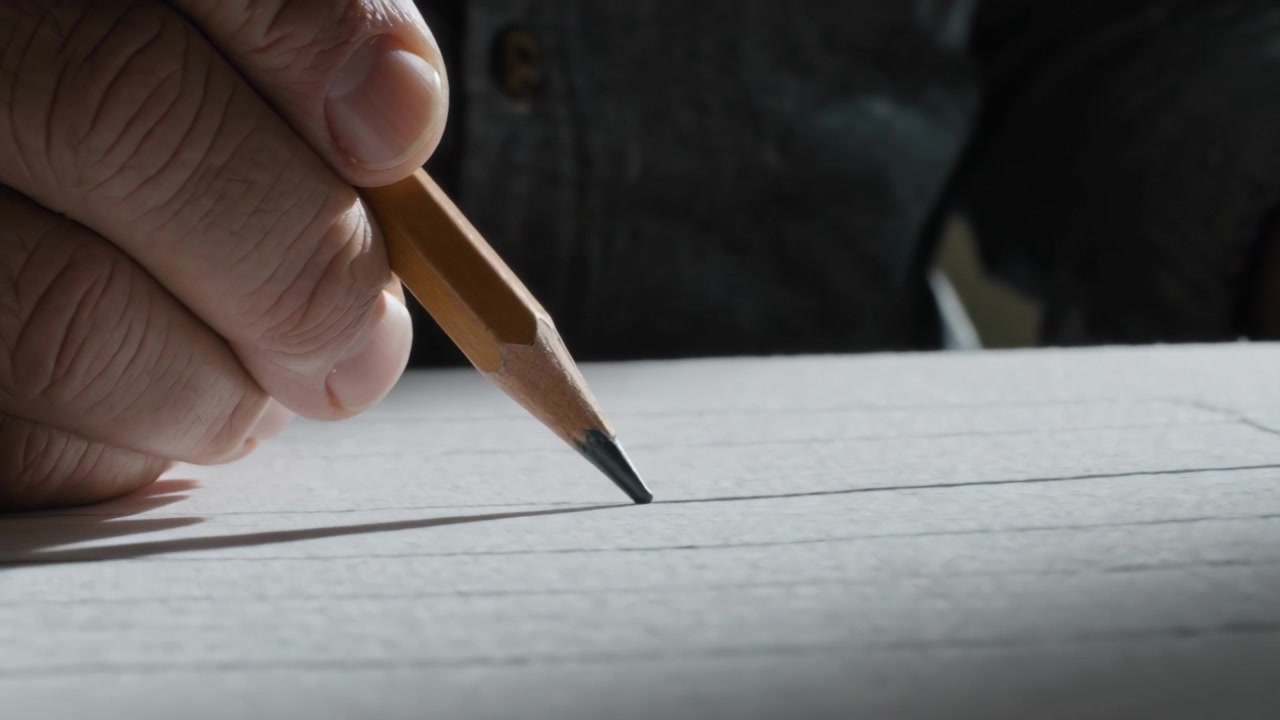 Touching pencil tip, right hand drawing lines on white paper at desk, drafting layout, denim shirt