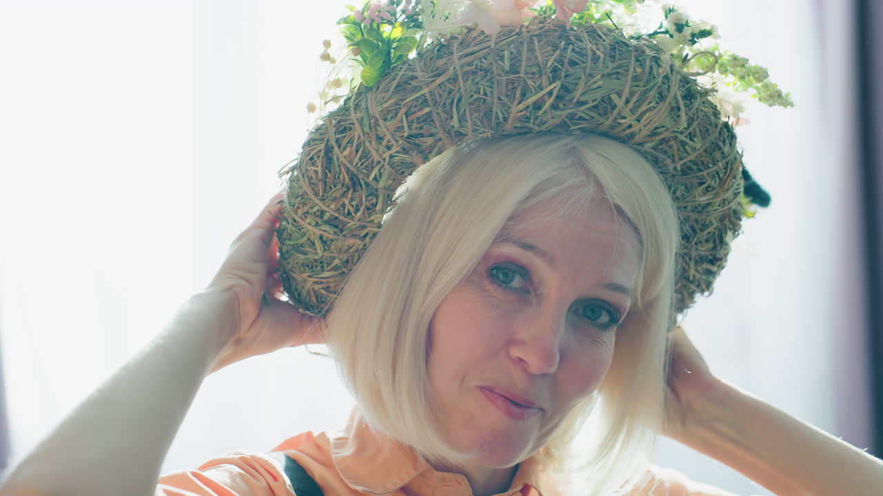 Smiling woman with blonde hair wearing rustic floral wreath hat made of hay and flowers, dressed in orange shirt with black overalls, adjusting accessory while standing indoors with natural sunlight