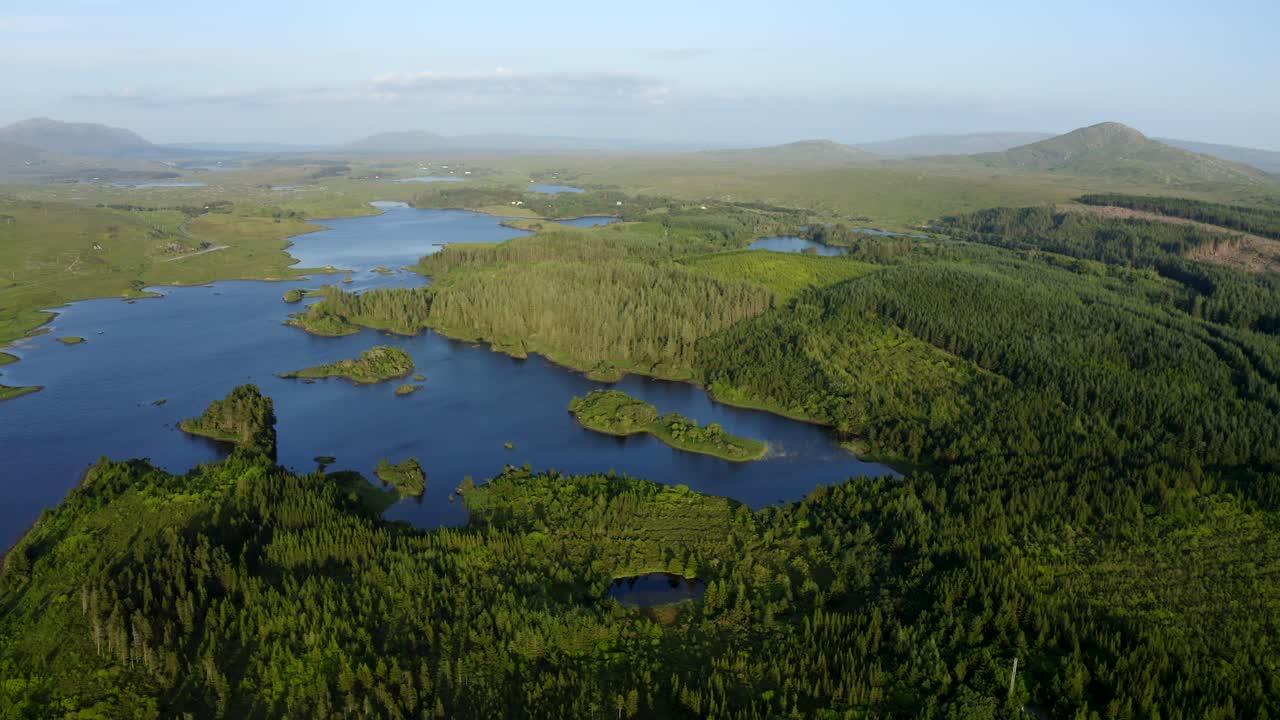 Ballynahinch Lake, Recess, Connemara, County Galway, Ireland, July 2021. Drone slowly tracks south over woods and islands with the Garraun mountain complex, Ballinafad and Maam in the distance