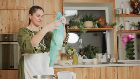 woman in green outfit stands in rustic kitchen holding blue piping bag preparing to bake with calm expression, surrounded by wooden cabinets, potted plants, oven, and countertop utensils
