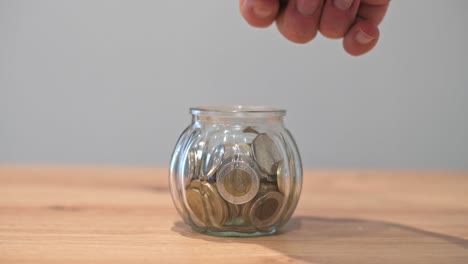 A close-up of a hand dropping a coin into a glass jar filled with coins, symbolizing saving money, financial growth, or budgeting.