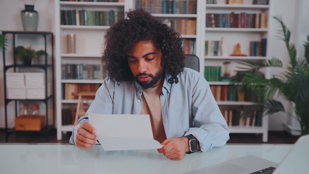 Young arabian man entrepreneur holds sheet of paper sits at table with laptop